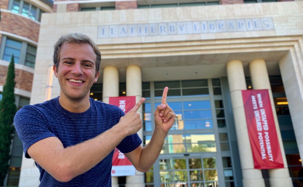 A young man in a blue t-shirt stands outside the front of the Leatherby Libraries, pointing with both his index fingers at the name on the front of the library