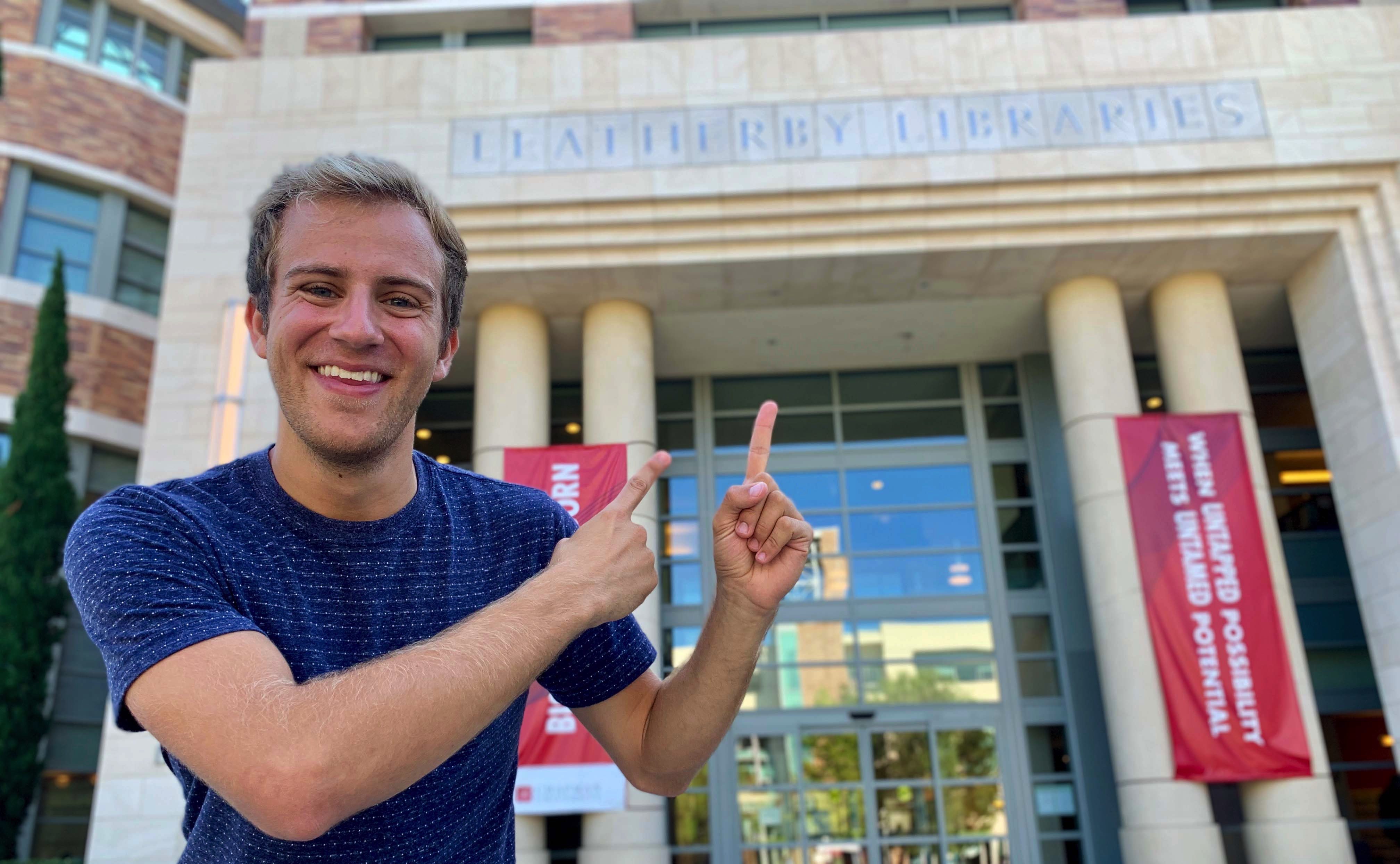 A young man stands in the front left of the frame, smiling and pointing with both hands at the words "LEATHERBY LIBRARIES" on the building behind him