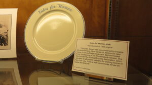 A white plate with the words "Votes for Women" in blue displayed next to explanatory text
