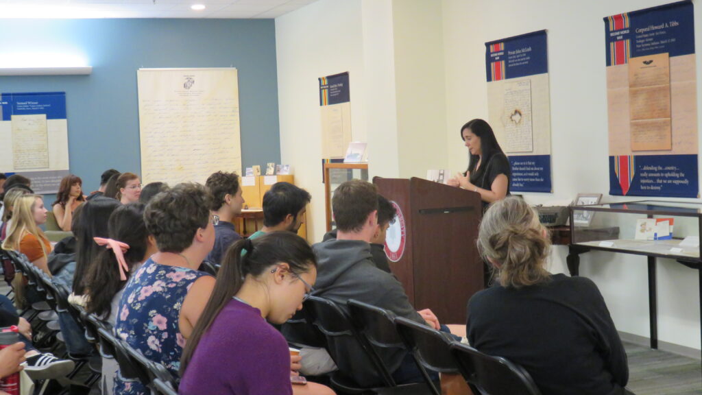 A crowd of people in the foreground sit listening to a woman speaking at a podium in the righthand background of the image