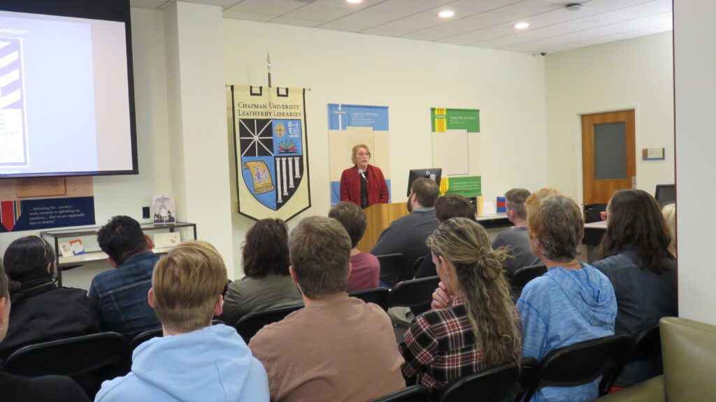 A woman in red stands in the background while a seated audience, photographed from behind, listens to her talk.