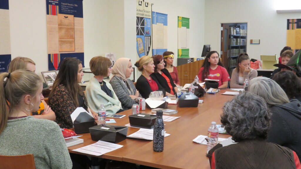 A group of women sit around a table, looking at a person speaking in the upper right corner of the image