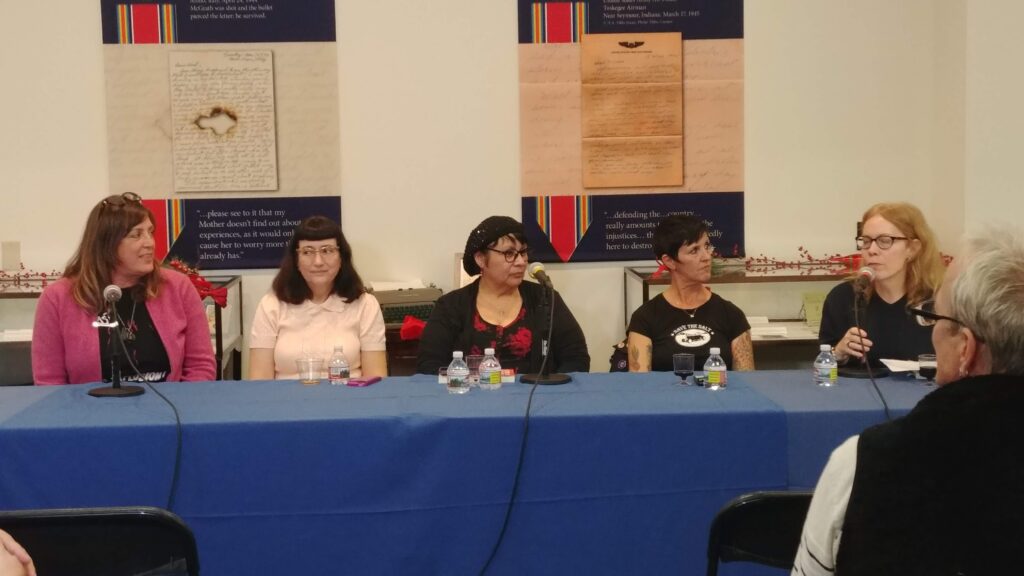 Five women sit at a table covered in a blue tablecloth, with microphones in front of them