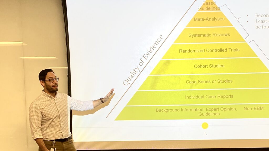 Ivan Portillo stands next to a projector screen displaying a yellow pyramid, which he is pointing at and explaining
