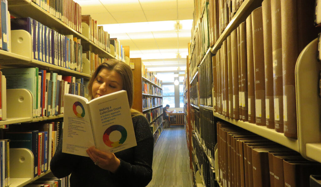 Kristin stands among shelves of books, reading an open book called Making a Collection Count