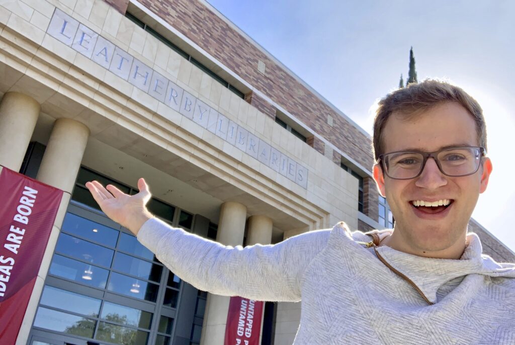 Jimmy Elinski stands smiling for the camera pointing at the entrance to the Leatherby Libraries behind him