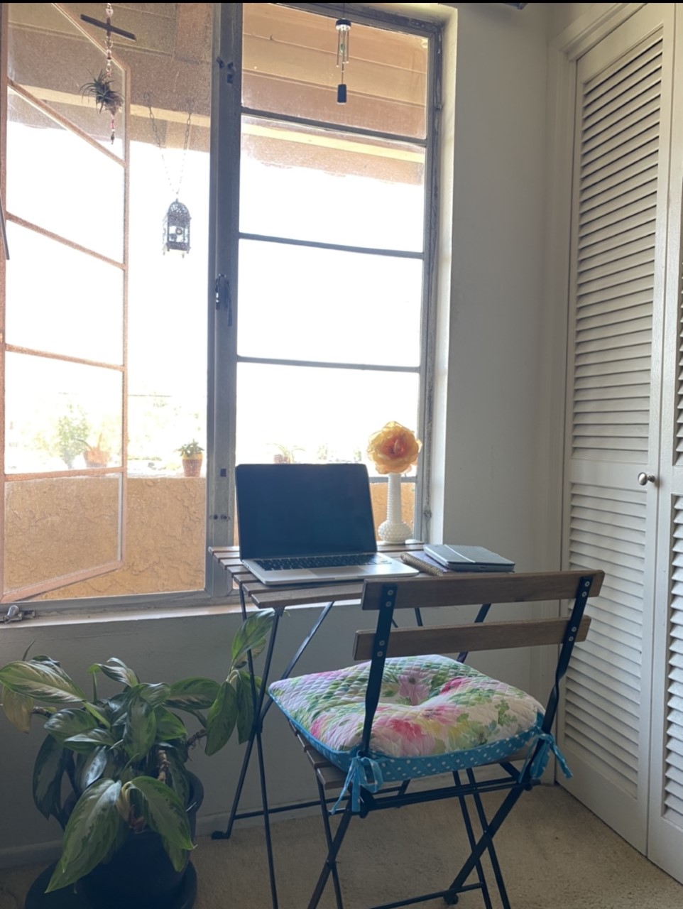 Photograph of a laptop on a table facing a large, open window, with a patio chair and cushion pulled up to the table