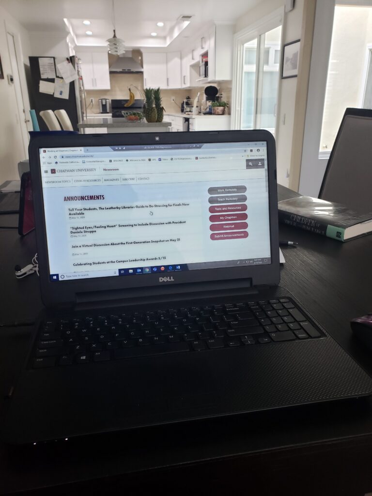 Photograph of a laptop on a dining room table, open to a browser window of the Chapman Newsroom. Visible in the background is a kitchen, and the book Children of Vengeance and Virtue sitting on the table