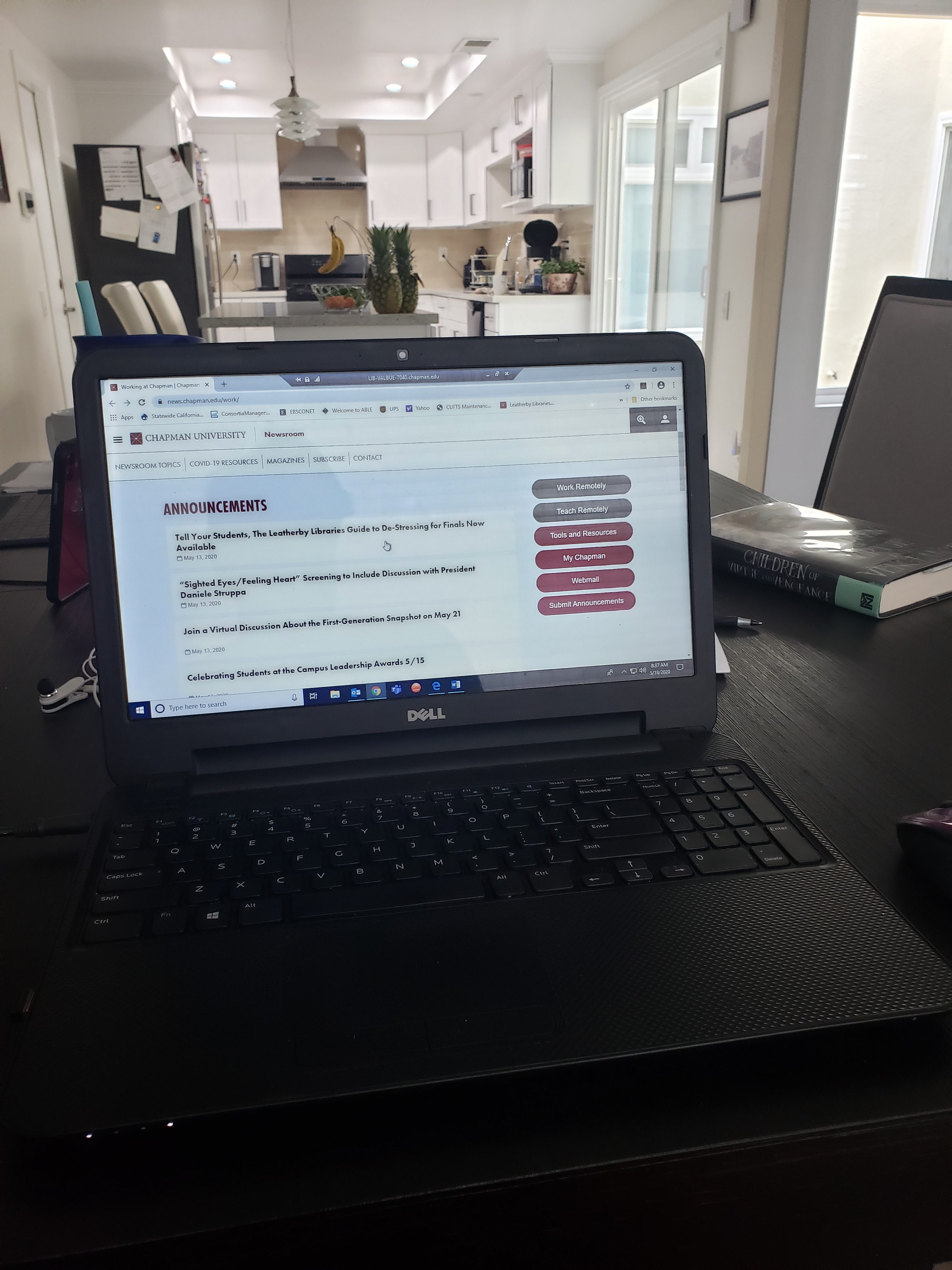 Photograph of a laptop on a dining room table, open to a browser window of the Chapman Newsroom. Visible in the background is a kitchen, and the book Children of Vengeance and Virtue sitting on the table