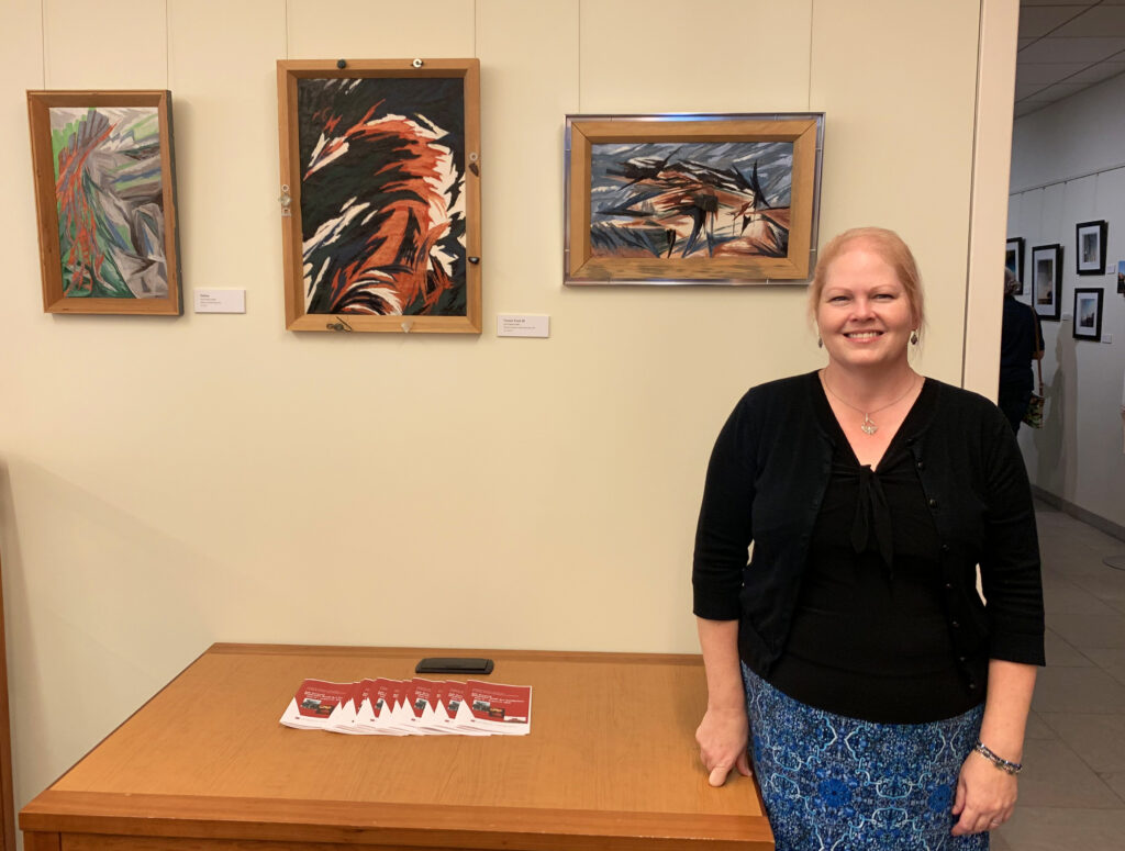 A woman stands next to a wooden table with three pieces of abstract art hanging on the wall to her right