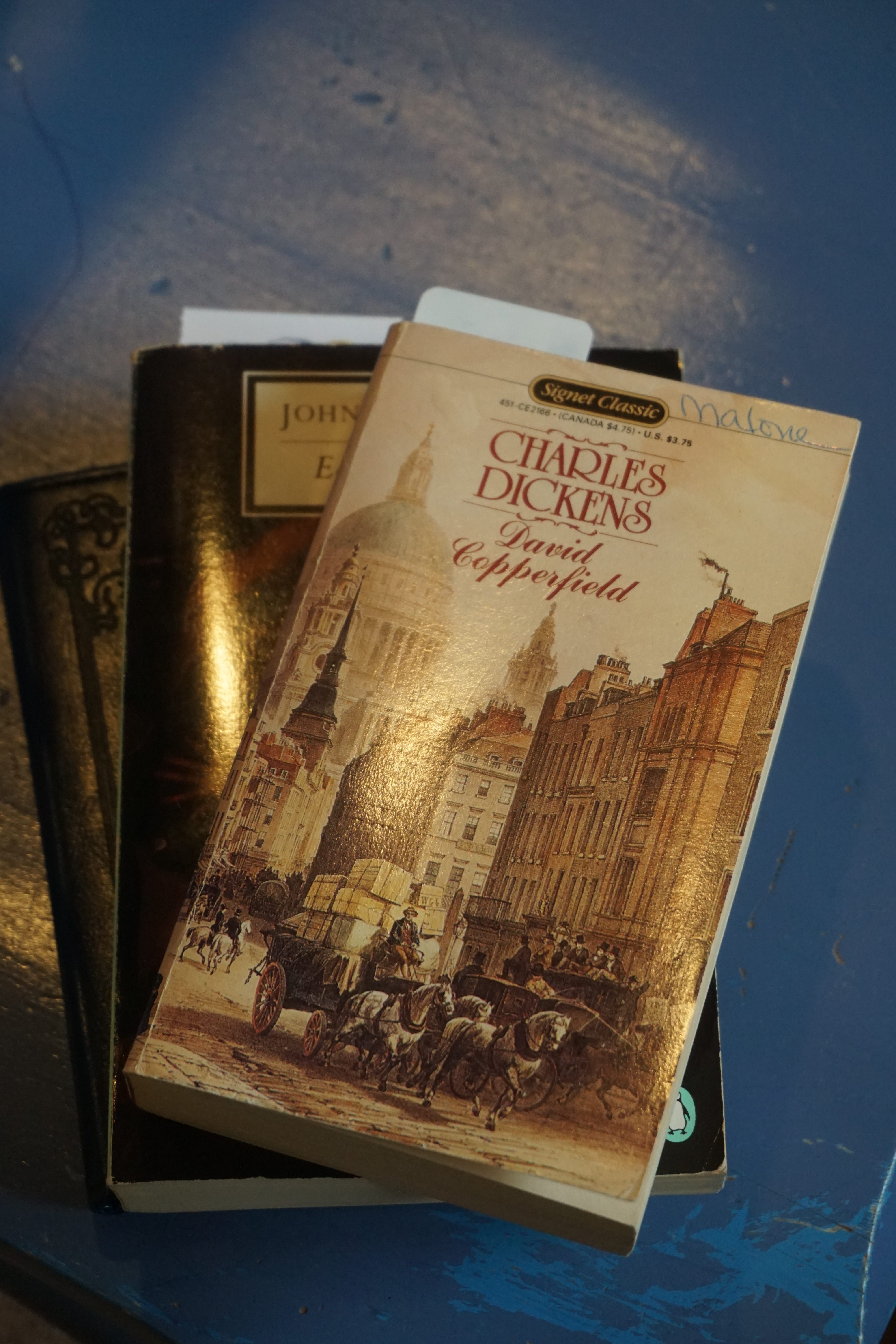 A stack of books on a blue background, shot from above. The top book is David Copperfield, by Charles Dickens. Below that is East of Eden, by John Steinbeck. On the bottom is a Kindle in a dark blue leather case