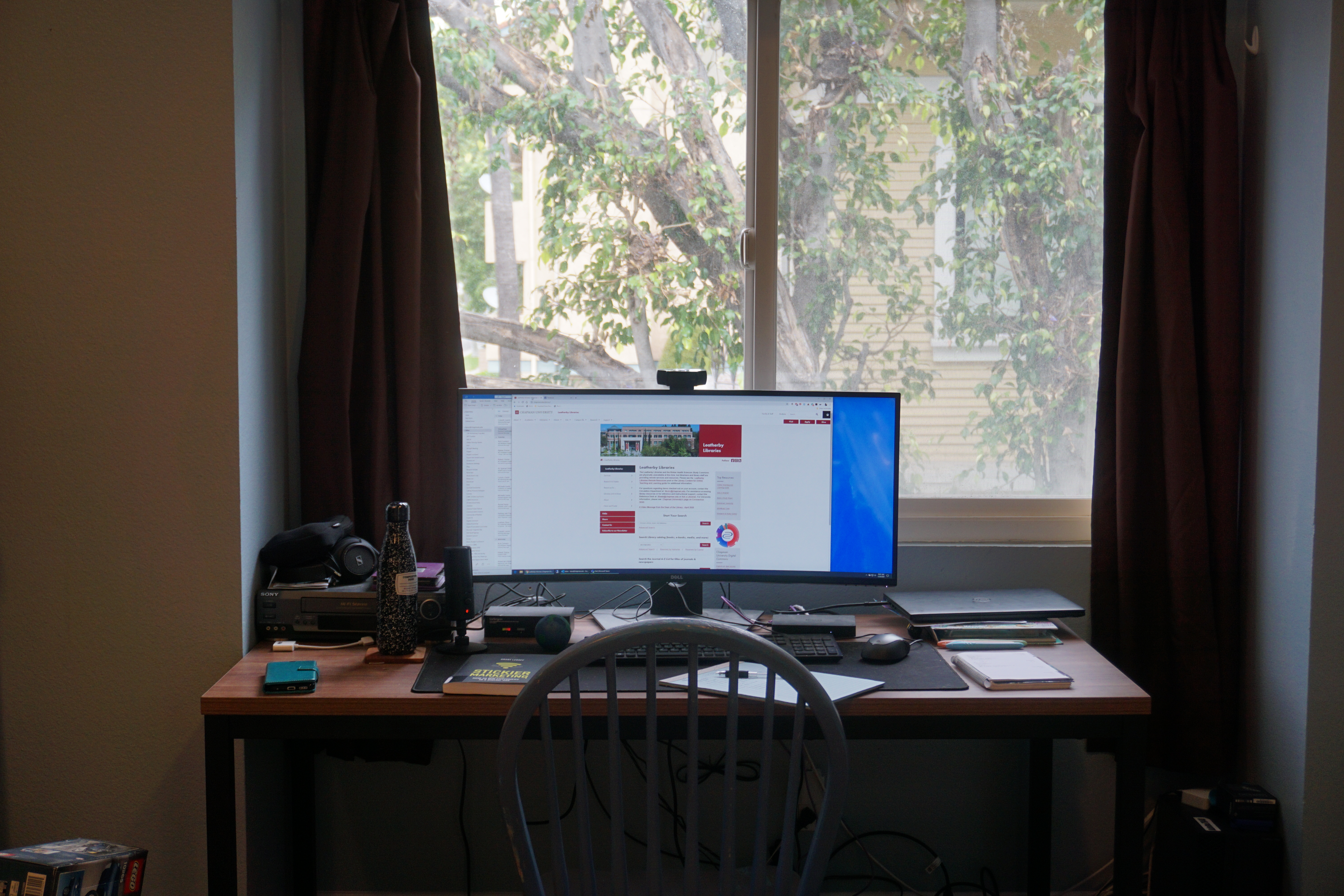 A desk in front of a window. On the desk are a large computer monitor open to the Leatherby Libraries home page, a water bottle, a phone, a keyboard, mouse, and two notebooks. Outside the window is a tree.