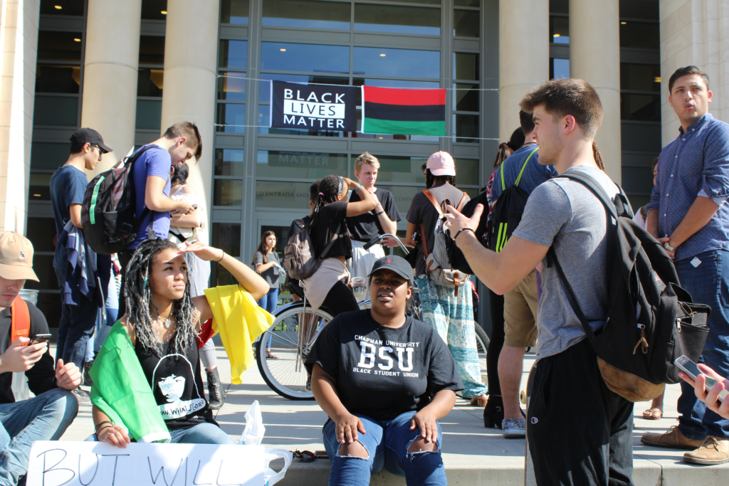 Photograph of the exterior of the Leatherby Libraries. A Black Lives Matter banner, along with a red, black, and green flag, is hanging over the door. Students sit and stand in front of the door, including two Black female students sitting talking to a standing White male student