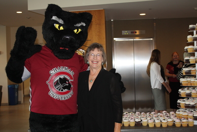 A person in a Pete the Panther mascot costume and a red Chapman University t-shirt poses with Dean Baldwin, who is wearing a black blouse, in front of a table filled with cupcakes, set in front of the elevator bay.