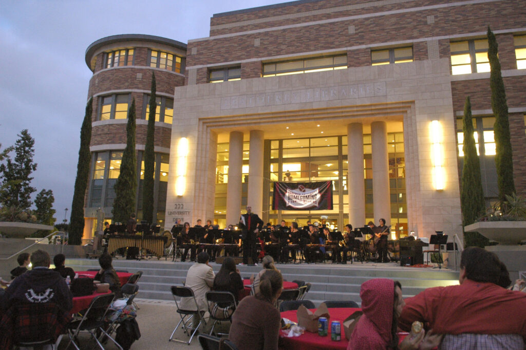 Exterior of the Leatherby Libraries at dusk with an orchestra playing on the stage, a Homecoming banner across the doors, and audience members seated at tables in the Attallah Piazza.