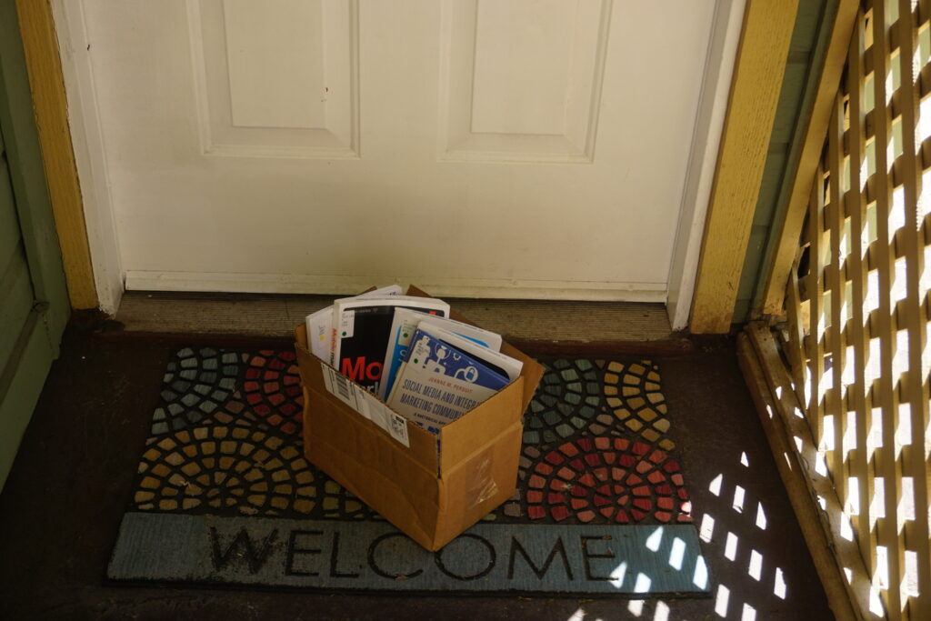 A cardboard box with library books standing up inside it sits on a Welcome mat in front of a door.