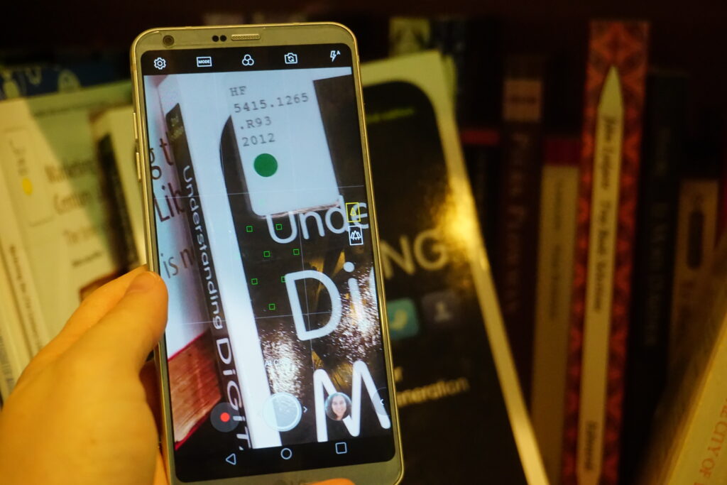 Close-up of a hand holding a cell phone, which is open to the camera feature and zoomed in on the library call number sticker on a book on a bookshelf.
