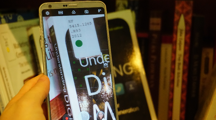 Close-up of a hand holding a cell phone, which is open to the camera feature and zoomed in on the library call number sticker on a book on a bookshelf.