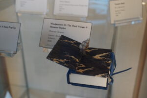 Close-up of a small blue book with a sailboat popping out. Behind it, a slightly out of focus information card reads, "Windjammers III: The Third Voyage: A Miniature Replica Duncan Gleason Lorson's Books Fullerton 2008"