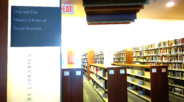 Rows of bookshelves covered in books. In the foreground is a wall with a sign that reads "Doy and Dee Henley Library of Social Sciences"
