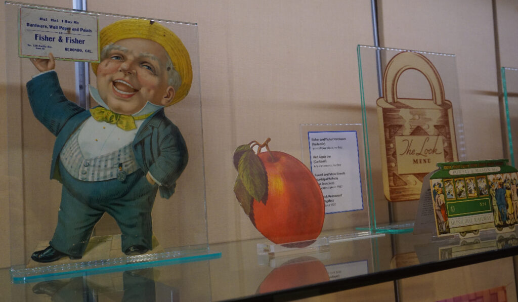 Several die-cut paper objects (a figure of a man yelling, an orange, and a covered wagon) on a glass display shelf.