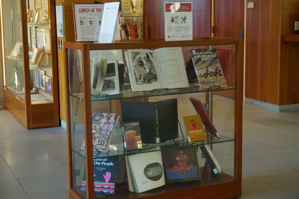 A glass display case filled with books on Native American art. Some books are closed to display their cover, and some are open to display an illustration inside.