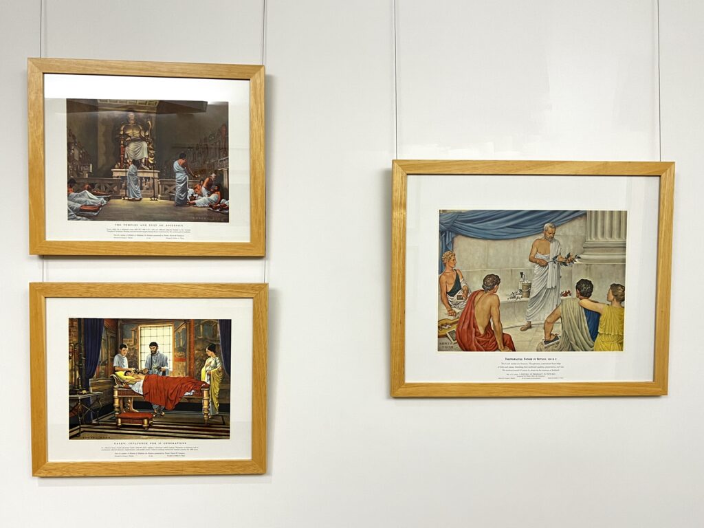 Three paintings of medical scenes framed and hanging on a white wall