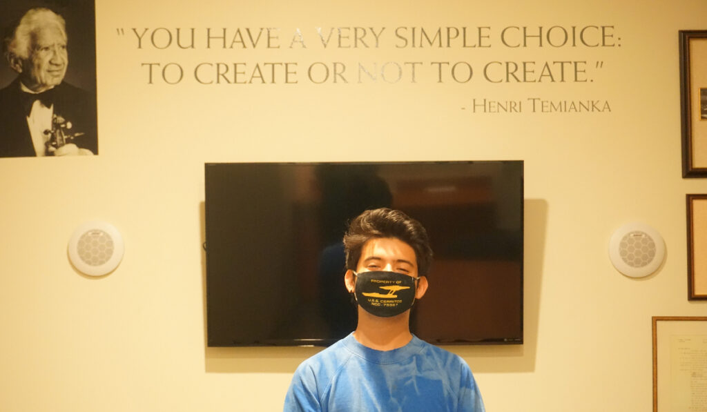 A young man wearing a face mask stands in front of a flat-screen television on a wall. Over the television are vinyl letters on the wall that read, "You have a very simple choice: to create or not to create. - Henri Temianka." In the upper left hand corner of the image is a portrait of Henri Temianka on the wall.