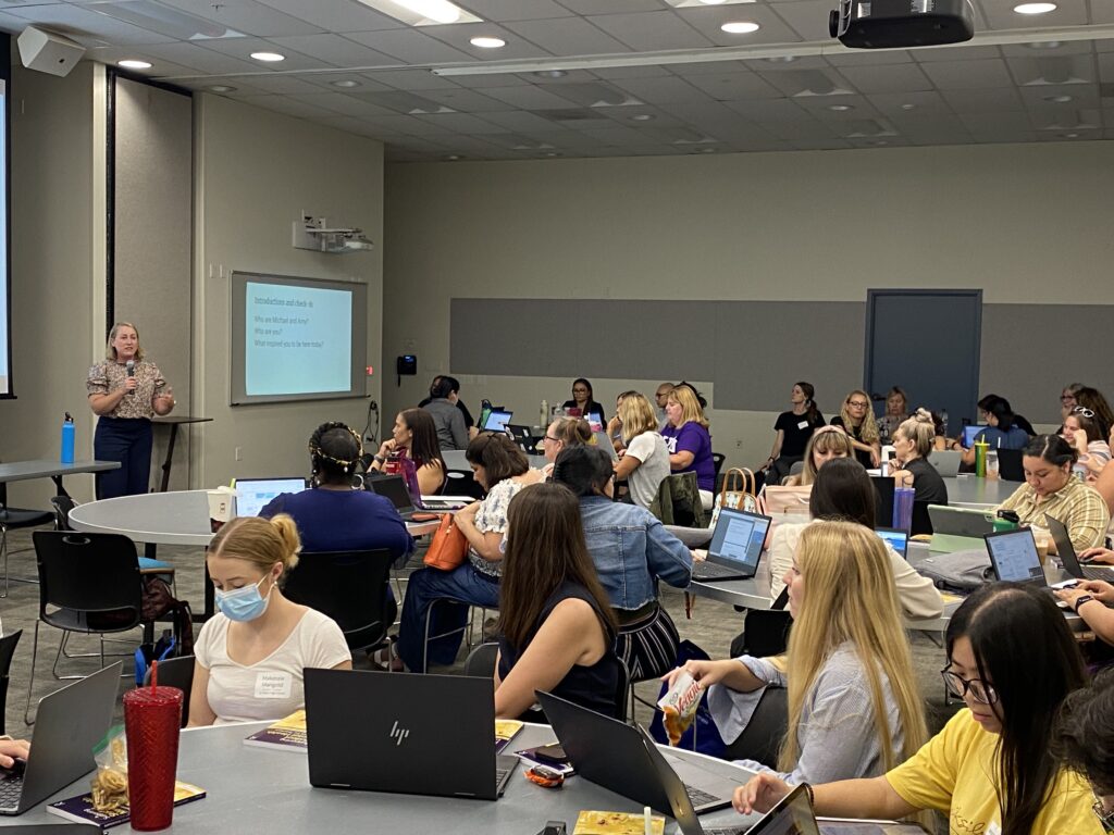 A professor is presenting in front of a group of educators sitting at round tables in a conference room.