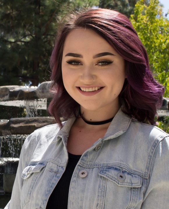 smiling young Caucasian female with shoulder length purple hair in light blue denim jacket with black t-shirt in front of a fountain
