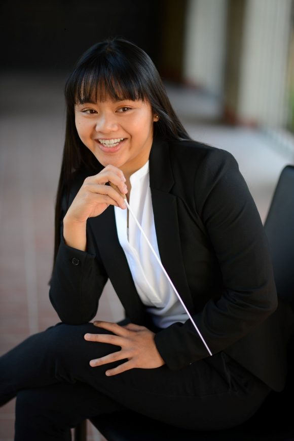 smiling Asian female in black suit seated and leaning forward with music baton in hand