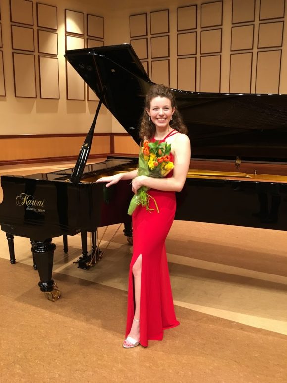 young smiling Caucasian woman in floor length sleeveless gown holding flower bouquet next to grand piano