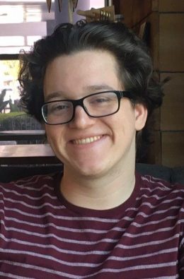 smiling young Caucasian male with short dark hair and black-rimmed glasses in burgundy striped shirt