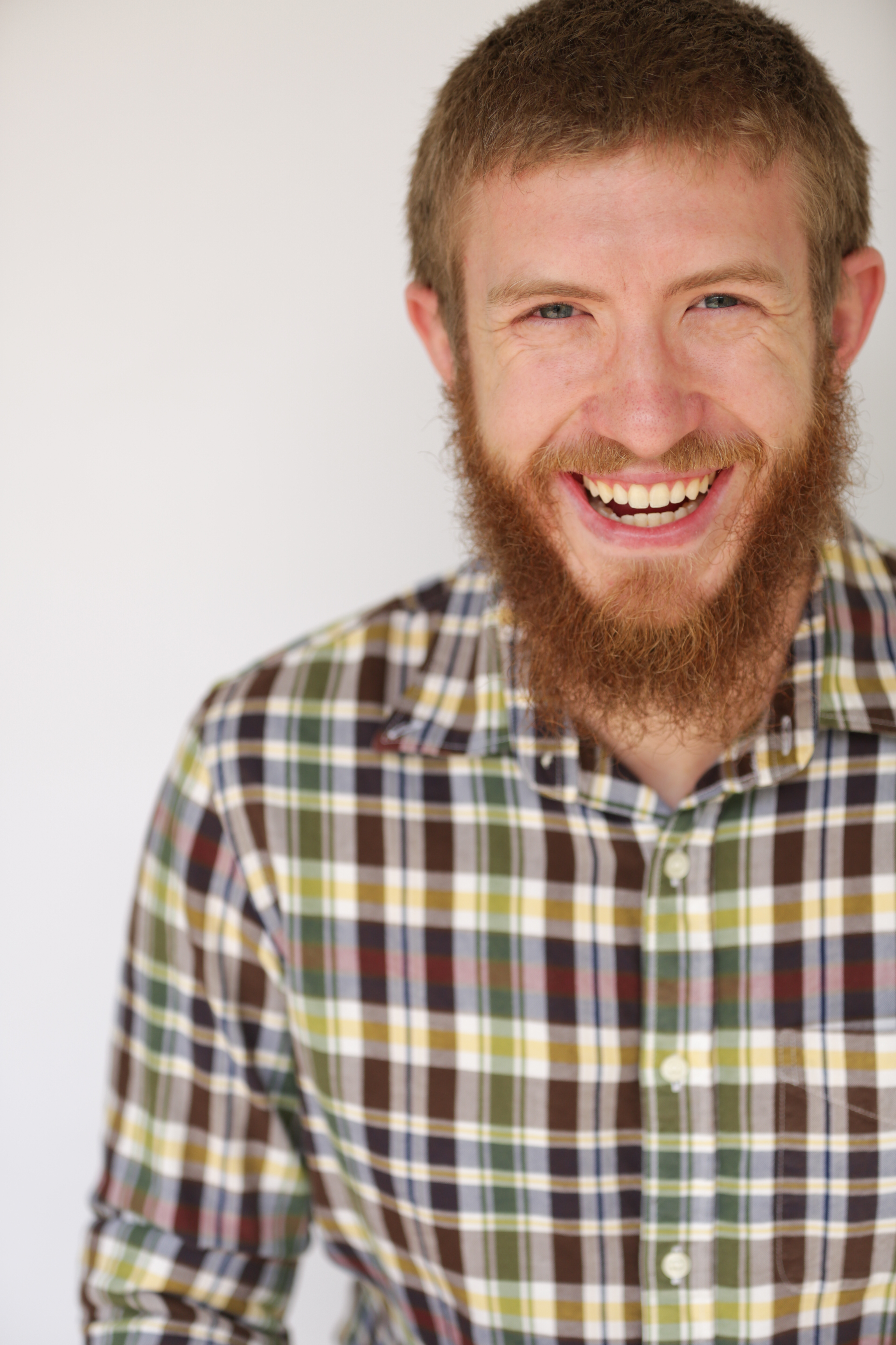 Smiling Caucasian man with curly red hair and beard wearing a blue and yellow plaid shirt.