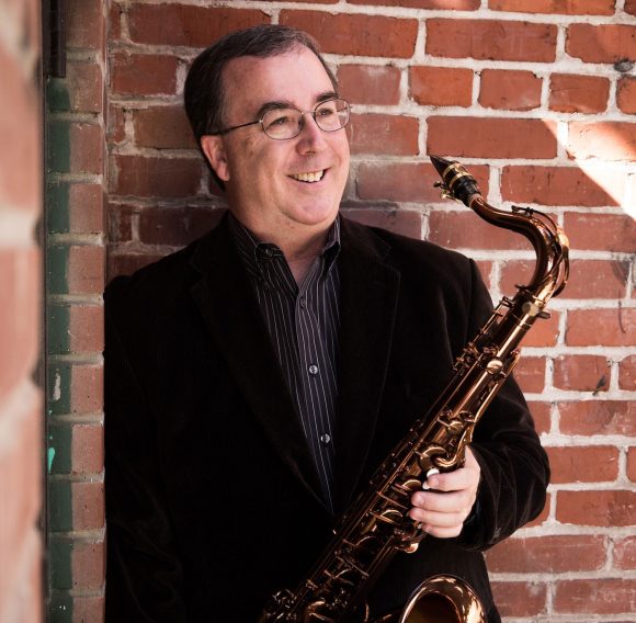 Smiling man in suit and glasses holding a saxophone.