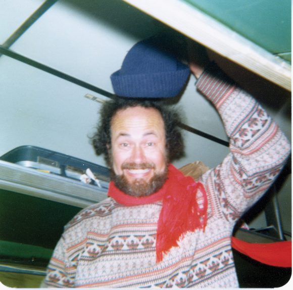 Smiling William hall with raised knit hat, sweater, and scarf while standing in a tour bus.