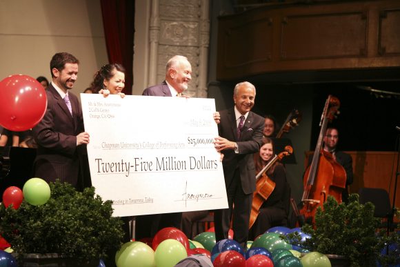 Four people on a stage holding a large replica check for $25M from an anonymous donor with balloons in the foreground and musicians in the background.