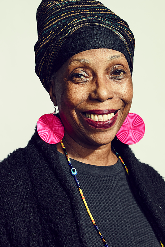 Smiling Black woman with traditional headdress, large pink disc earrings, and black cardigan.