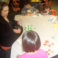 Woman sits at table with children, playing a game.