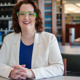 Portrait photograph of alumnus Jodee Storm Sullivan seated in the Fowler School of Law library, wearing a white blazer, blue shirt and distinctive green glasses.