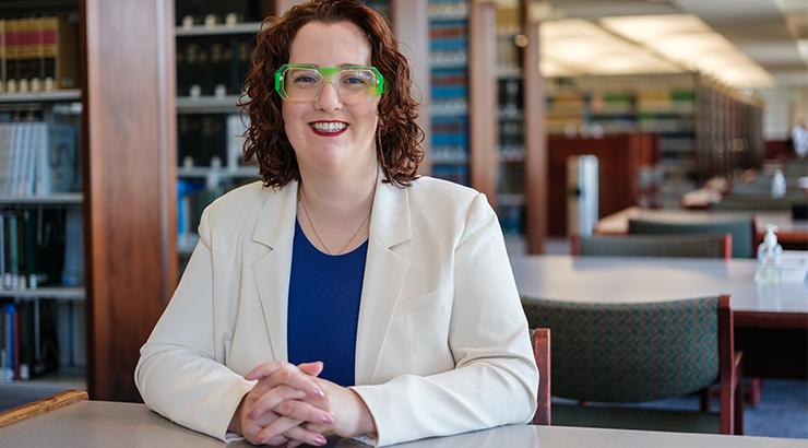 Portrait photograph of alumnus Jodee Storm Sullivan seated in the Fowler School of Law library, wearing a white blazer, blue shirt and distinctive green glasses.