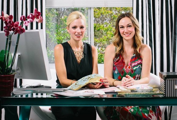 Two women, Naomi and her partner, sitting at a desk in their office smiling and looking at the camera