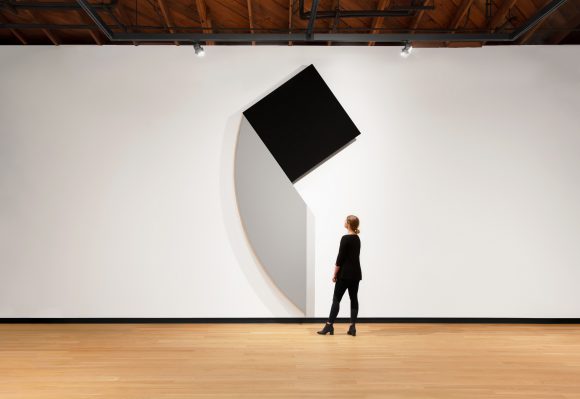 Image of woman dressed in black standing in front of a triangular-shaped large painting. The painting is black and grey and hung on a white wall.