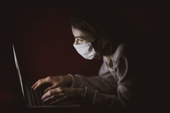 Woman typing on a laptop in the dark while wearing a mouth and nose mask