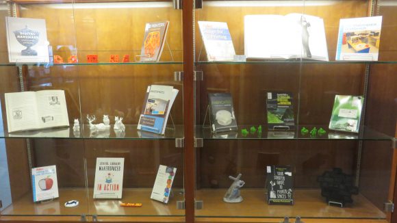 A display case with six glass shelves, each holding between one and three books and between two and six small 3D-printed objects