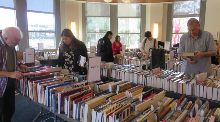 Several adults, some in the foreground and some in the back, look through tables of books, organized by genre.
