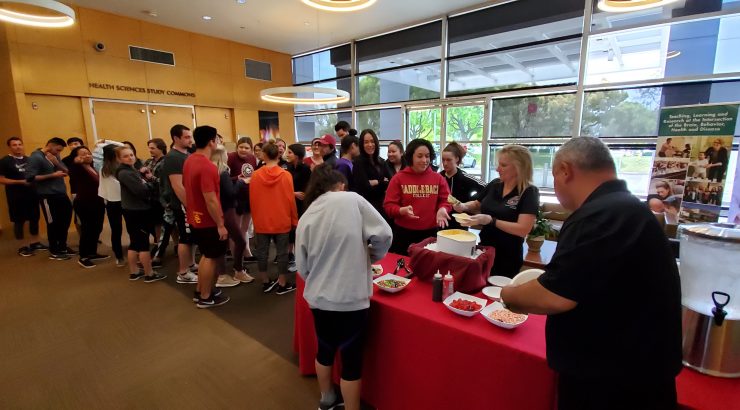 A long line of students, moving from the center to the top left-hand corner of the image, waits for ice cream to be served from a red table in the lower right-hand corner.