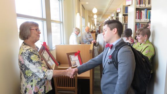 A woman on the left shakes the hand of a young male college student on the right, handing him a wrapped, framed photo, while people look on from the background.