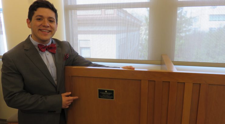 A young man stands next to a study carrel, pointing at the plaque on it that lists his name.
