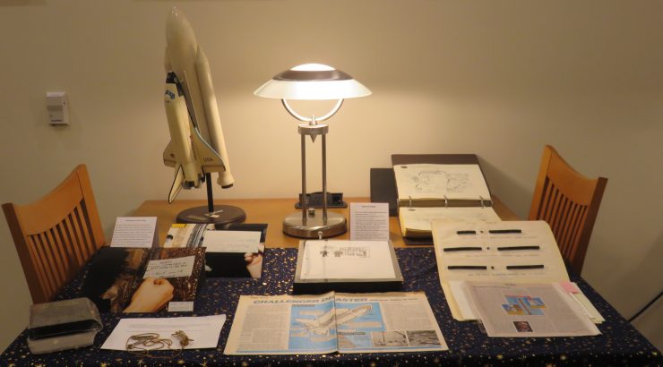 Multiple materials, some printed, some photographs, and one model of the Challenger shuttle, displayed on a table with a lamp in the background.